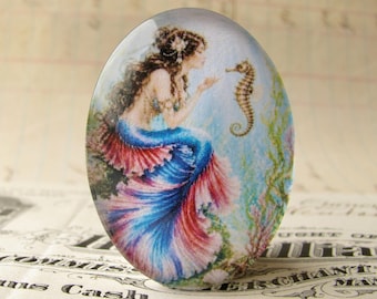 May include: Oval-shaped glass cabochon featuring a mermaid with long brown hair, a blue top, and a flowing blue and red skirt. The mermaid is interacting with a seahorse against a light blue and green background.