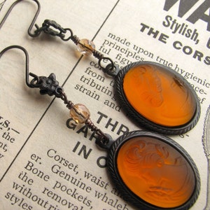 May include: A pair of earrings with amber glass cameo pendants featuring a woman's profile. The earrings have a bronze finish and are accented with small amber glass beads.