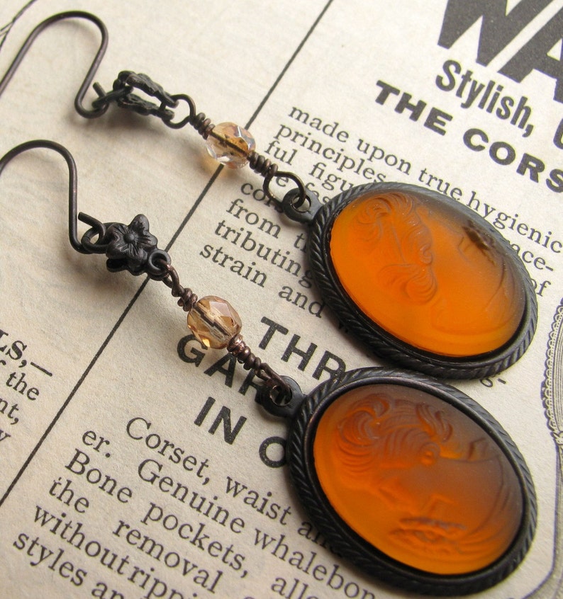 May include: A pair of earrings with amber glass cameo pendants featuring a woman's profile. The earrings have a bronze finish and are accented with small amber glass beads.