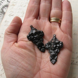 May include: Two black metal charms featuring a woman's face with ornate details. The charms are in the shape of a teardrop and have a loop at the top for attaching to a necklace or bracelet.