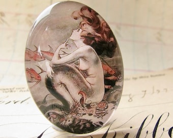 May include: Oval-shaped glass cabochon featuring a vintage illustration of a mermaid with flowing brown hair, surrounded by fish. The artwork is in shades of gray, red, and white, with a glossy finish. The cabochon is likely for jewelry or craft projects.