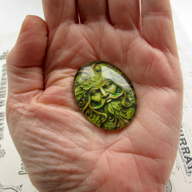 Op de afbeelding: Een groene glazen cabochon met een gedetailleerd beeld van de Groene Man, een mythische figuur die vaak wordt geassocieerd met natuur en vruchtbaarheid.