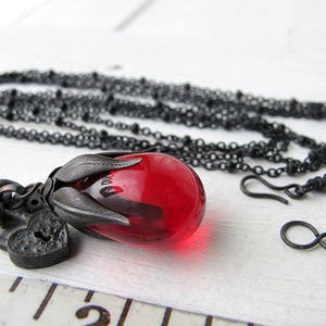Op de afbeelding: Een zwarte ketting met een hanger met een grote, rode, druppelvormige glazen kraal. De hanger is gevat in een donkere metalen setting met bladachtige details en een kleine hartvormige bedel. De ketting is ook zwart.