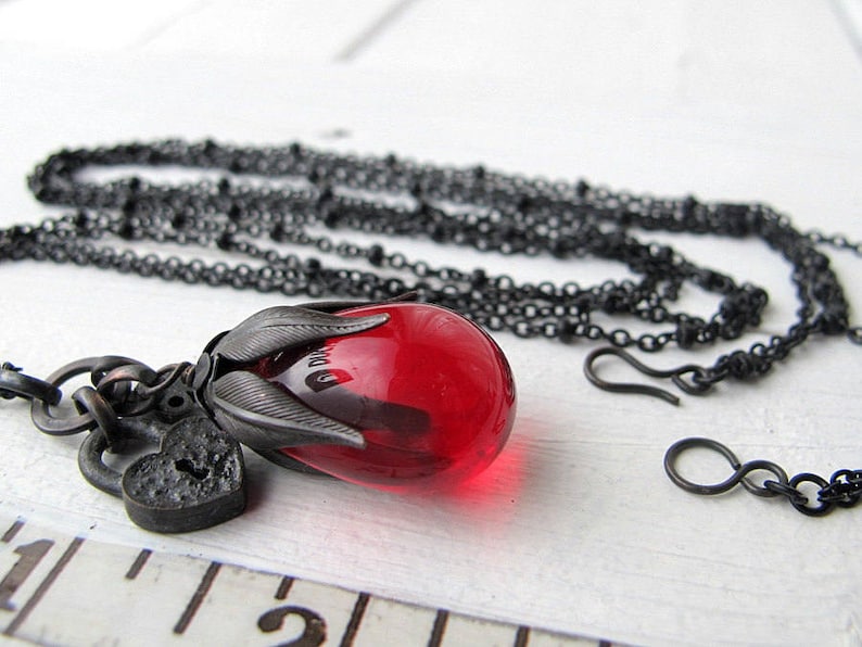 Op de afbeelding: Een zwarte ketting met een hanger met een grote, rode, druppelvormige glazen kraal. De hanger is gevat in een donkere metalen setting met bladachtige details en een kleine hartvormige bedel. De ketting is ook zwart.