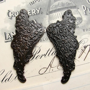 May include: Two ornate metal wings with a dark bronze finish. The wings have a detailed floral design and are perfect for crafting projects.