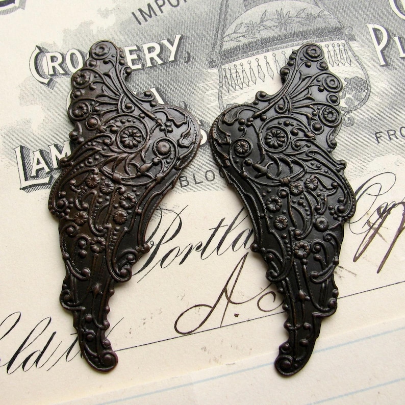 May include: Two ornate metal wings with a dark bronze finish. The wings have a detailed floral design and are perfect for crafting projects.