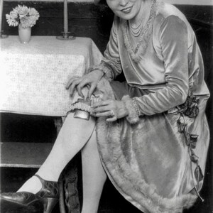 May include: A woman wearing a light-coloured dress and a hat with a flower on it. She is sitting at a table with a vase of flowers on it. She is holding a small object in her hand.