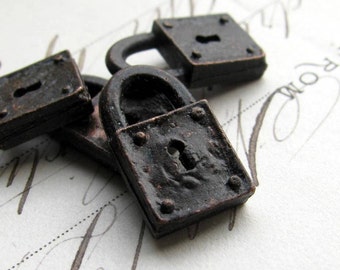 May include: Close-up of several small, antique-style padlocks. The padlocks are rectangular with a dark, weathered finish, featuring keyholes and rounded handles. The metal has a textured appearance, suggesting age and use.