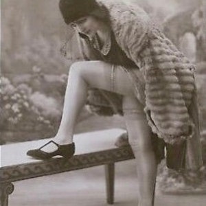 May include: A woman in a fur coat and stockings is standing on a bench, adjusting her shoe. She is wearing a black hat and a black dress. The photograph is in black and white.