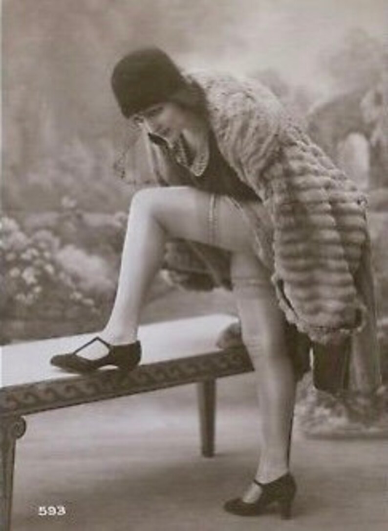 May include: A woman in a fur coat and stockings is standing on a bench, adjusting her shoe. She is wearing a black hat and a black dress. The photograph is in black and white.