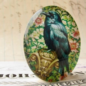 May include: A round glass cabochon with a vintage image of a blue raven perched on a gold ornate pedestal. The raven is surrounded by a floral pattern with pink and white flowers.