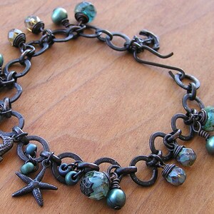 K&ouml;nnte beinhalten: Ein handgefertigtes Armband mit einer dunklen Metallkette und verschiedenen Anh&auml;ngern. Die Anh&auml;nger umfassen ein Seepferdchen, einen Seestern und mehrere facettierte Perlen in T&uuml;rkis-, Gr&uuml;n- und Klart&ouml;nen. Das Armband hat auch kleine, runde, t&uuml;rkisfarbene Perlen.
