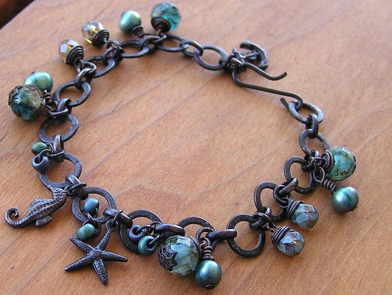 K&ouml;nnte beinhalten: Ein handgefertigtes Armband mit einer dunklen Metallkette und verschiedenen Anh&auml;ngern. Die Anh&auml;nger umfassen ein Seepferdchen, einen Seestern und mehrere facettierte Perlen in T&uuml;rkis-, Gr&uuml;n- und Klart&ouml;nen. Das Armband hat auch kleine, runde, t&uuml;rkisfarbene Perlen.