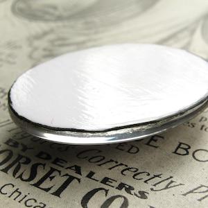 Op de afbeelding: Een ronde, witte metalen knoop met een zilveren rand. De knoop heeft een glad, licht getextureerd oppervlak.