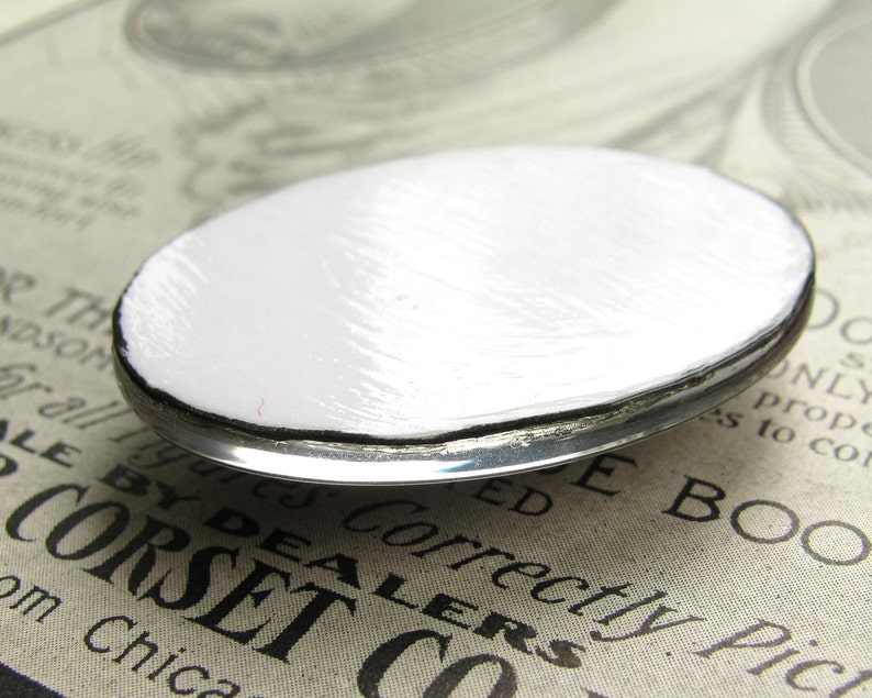 Op de afbeelding: Een ronde, witte metalen knoop met een zilveren rand. De knoop heeft een glad, licht getextureerd oppervlak.