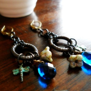 Puede incluir: Un par de pendientes colgantes con cuentas de cristal azul, flores de porcelana blanca y un dije de lib&eacute;lula. Los pendientes est&aacute;n hechos con una combinaci&oacute;n de metal dorado y plateado.