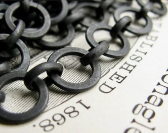 May include: Close-up of a dark gray metal chain with circular links. The chain is lying on a vintage paper with the text "ESTABLISHED 1868" visible. The chain's texture and the paper's aged appearance create a rustic aesthetic.