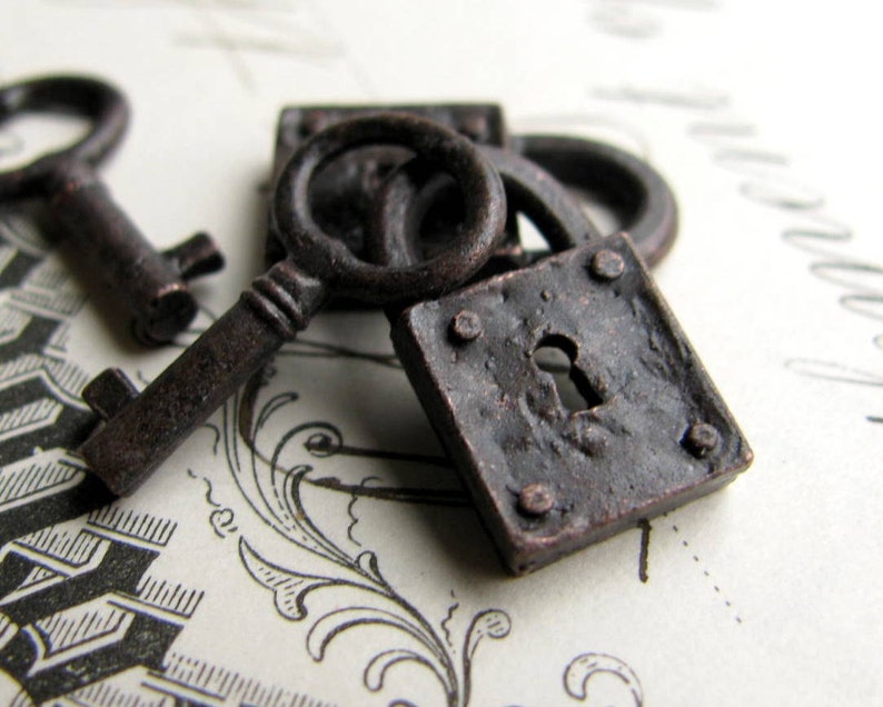 K&ouml;nnte beinhalten: Nahaufnahme von antiken, dunkelbraunen Metallschl&uuml;sseln und einem kleinen Vorh&auml;ngeschloss. Die Schl&uuml;ssel weisen komplizierte Details auf und sind in der N&auml;he des Schlosses platziert, was auf ein Vintage- oder historisches Thema hindeutet. Das Schloss ist quadratisch mit einem Schl&uuml;sselloch.