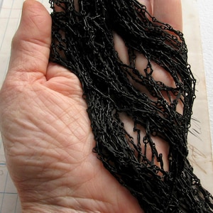 May include: A long, matte black chain necklace featuring a series of interconnected, elongated oval links. The chain is composed of multiple strands of dark, uniform metal links that create a dense, textured appearance across the entire length.