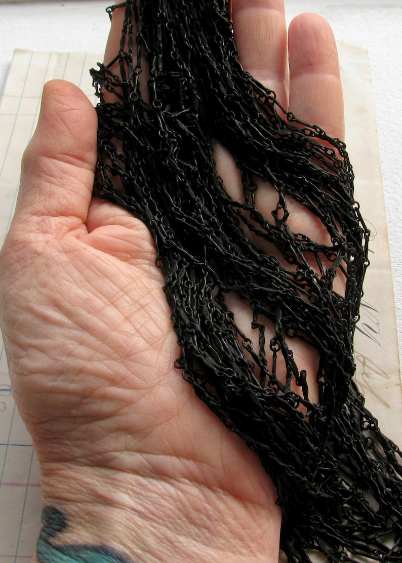 May include: A long, matte black chain necklace featuring a series of interconnected, elongated oval links. The chain is composed of multiple strands of dark, uniform metal links that create a dense, textured appearance across the entire length.