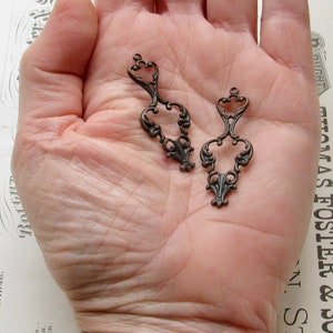 May include: Two antique bronze metal filigree charms with a decorative design. The charms are in the shape of a heart with a loop at the top.