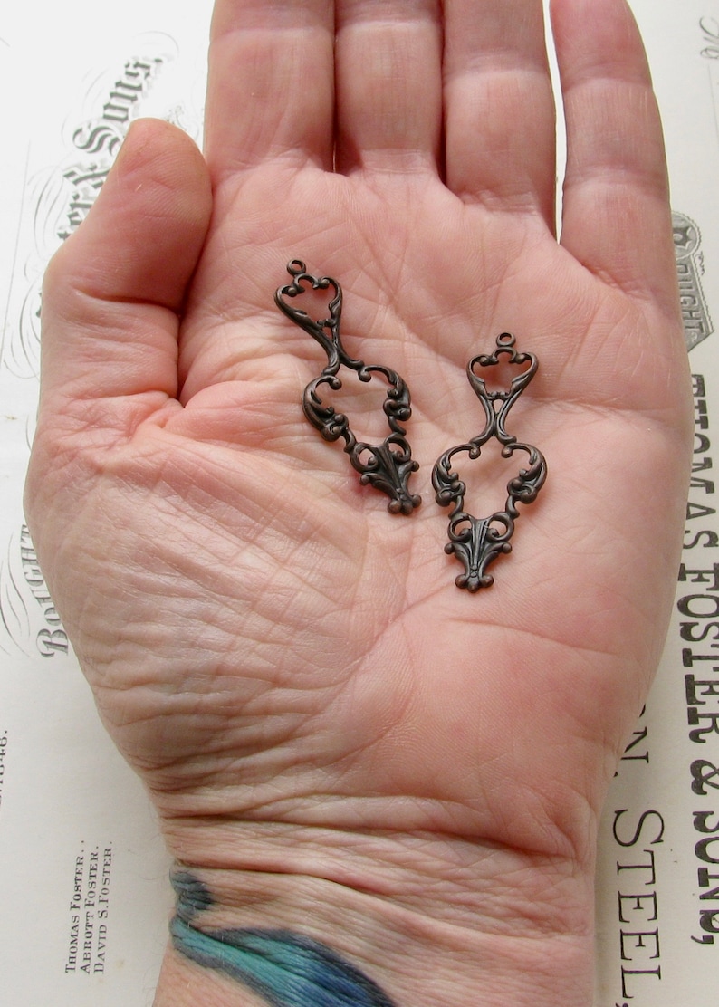 May include: Two antique bronze metal filigree charms with a decorative design. The charms are in the shape of a heart with a loop at the top.