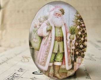 May include: An oval glass cabochon featuring a vintage-style illustration of Santa Claus wearing a pale pink coat with white fur trim over a green suit, carrying a green sack and standing beside a decorated Christmas tree.