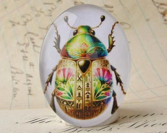 May include: An oval-shaped glass paperweight featuring a colorful, detailed illustration of a scarab beetle. The beetle's shell is adorned with intricate patterns in gold, green, blue, and pink, with floral and hieroglyphic elements. The paperweight is set against a blurred background.