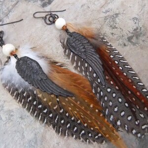 May include: A pair of feather earrings with a black metal wing charm and a white bead. The feathers are brown, orange, and gray with white spots.