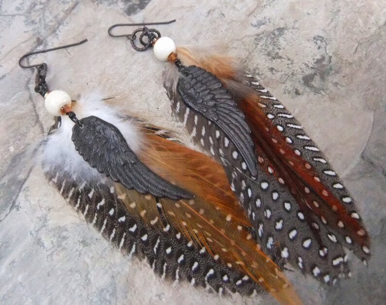 May include: A pair of feather earrings with a black metal wing charm and a white bead. The feathers are brown, orange, and gray with white spots.
