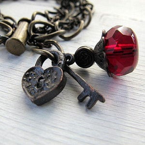 Op de afbeelding: Een vintage armband met een hartvormig slot, een sleutel en een facetgeslepen rode glazen kraal. De armband heeft een donkere, antiek bronzen afwerking, met een ketting en sluiting. De sieraden liggen op een wit houten oppervlak.