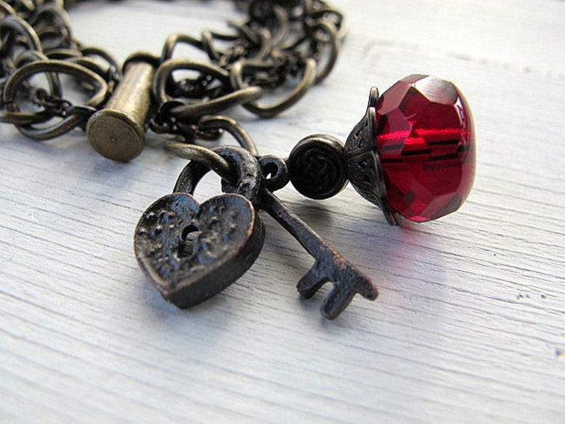 Op de afbeelding: Een vintage armband met een hartvormig slot, een sleutel en een facetgeslepen rode glazen kraal. De armband heeft een donkere, antiek bronzen afwerking, met een ketting en sluiting. De sieraden liggen op een wit houten oppervlak.