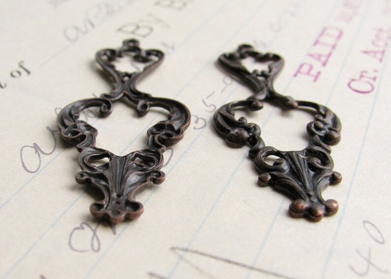 May include: Two ornate metal embellishments with a decorative, scroll-like design. The embellishments are a dark bronze color and have a vintage look.