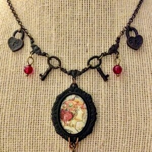 Op de afbeelding: Een bronskleurige ketting met een vintage esthetiek. De ketting heeft hartvormige slotbedels, sleutelbedels en een centrale ovale hanger met een vrouwenprofiel. Rode kralen bungelen aan de sleutels en de centrale hanger.