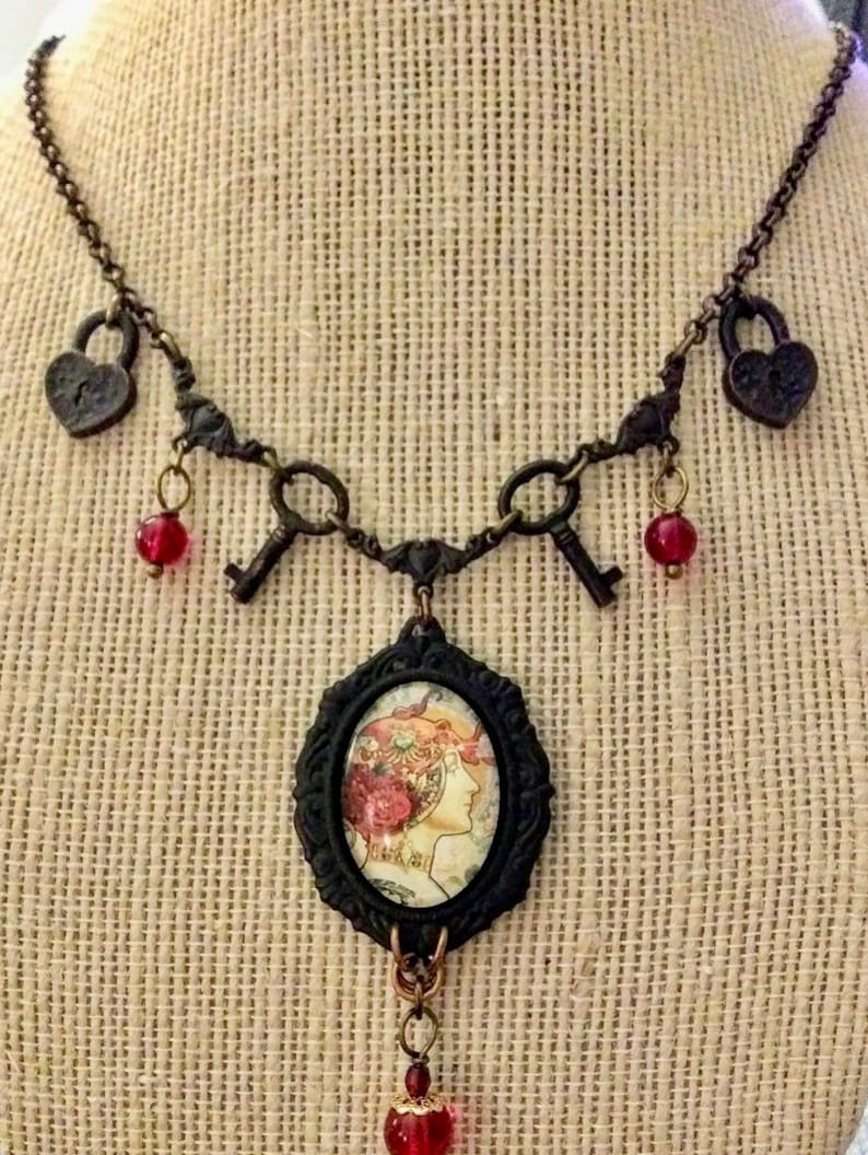 Op de afbeelding: Een bronskleurige ketting met een vintage esthetiek. De ketting heeft hartvormige slotbedels, sleutelbedels en een centrale ovale hanger met een vrouwenprofiel. Rode kralen bungelen aan de sleutels en de centrale hanger.