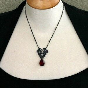 May include: A black necklace with a delicate, ornate pendant featuring a heart-shaped design and a dangling red teardrop-shaped crystal.