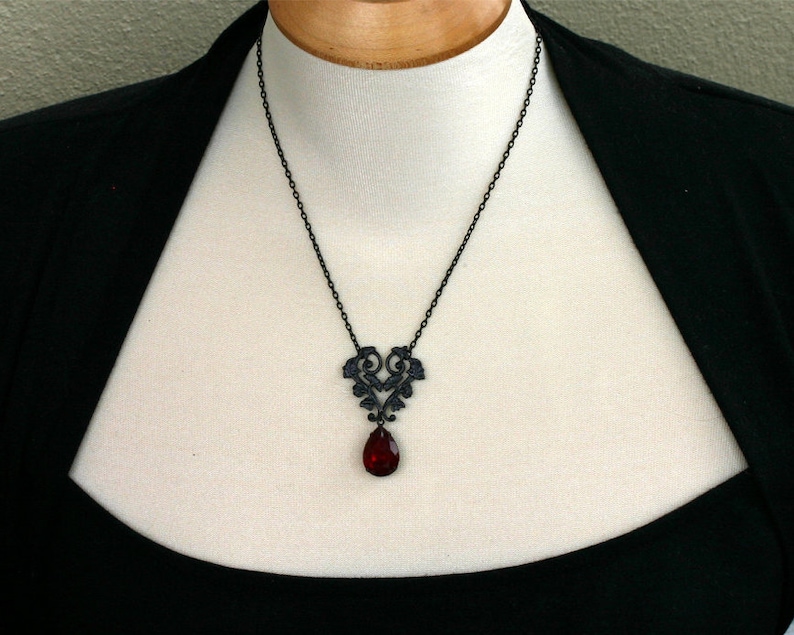 May include: A black necklace with a delicate, ornate pendant featuring a heart-shaped design and a dangling red teardrop-shaped crystal.