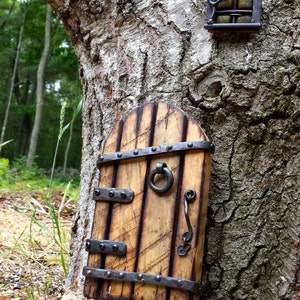 Op de afbeelding: Een miniatuur houten elfendeur met metalen hardware, inclusief een handvat en scharnieren, is op een boomstam gemonteerd. Een bijpassend raam bevindt zich boven de deur. De deur is lichtbruin met donkerbruine accenten.
