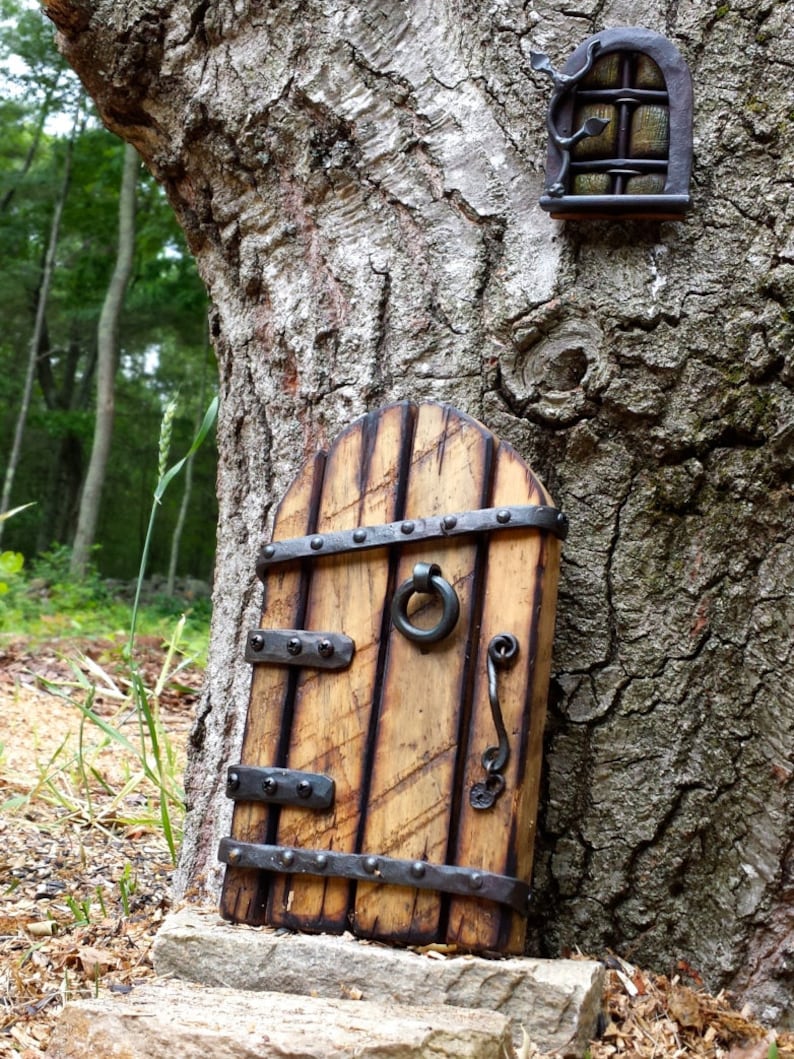 Op de afbeelding: Een miniatuur houten elfendeur met metalen hardware, inclusief een handvat en scharnieren, is op een boomstam gemonteerd. Een bijpassend raam bevindt zich boven de deur. De deur is lichtbruin met donkerbruine accenten.