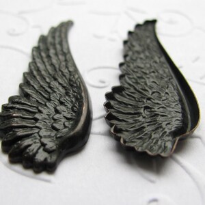 May include: Two black metal angel wings with a detailed feathered design. The wings are slightly curved and have a smooth, polished finish.