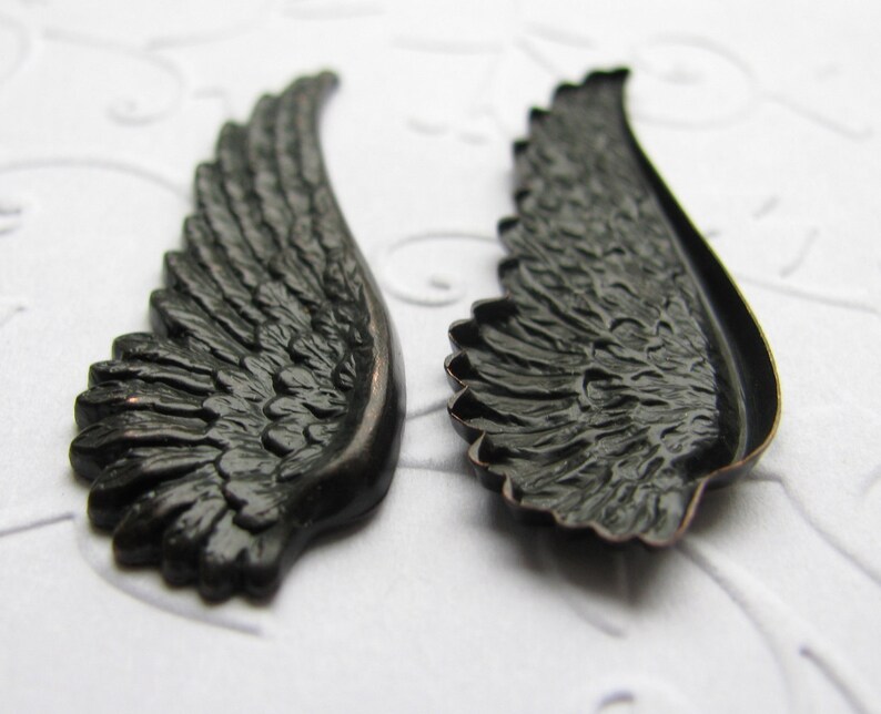 May include: Two black metal angel wings with a detailed feathered design. The wings are slightly curved and have a smooth, polished finish.