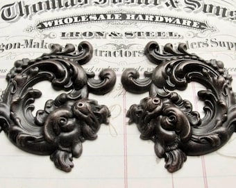 May include: Two ornate metal embellishments with a dark bronze finish. Each piece features a swirling, scroll-like design with a detailed face at the bottom.