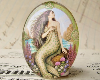 Op de afbeelding: Een ovale glazen cabochon met een kleurrijke illustratie van een zeemeermin met lang donker haar die op een rots bij de zee zit. De scène bevat paarse en witte wilde bloemen, een groene decoratieve rand en een blauwe oceaan.