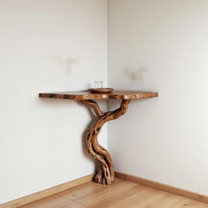 Op de afbeelding: Een handgemaakte houten hoektafel met een natuurlijke boomstamvoet. De tafel heeft een blad met levende rand. Een klein glas en een houten bordje staan op de tafel. De tafel is een decoratief accentstuk.