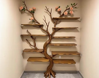 May include: A unique, tree-shaped wooden shelf unit with multiple shelves. The brown, sculpted trunk supports several horizontal shelves. Artificial pink roses and green leaves adorn the branches, adding a touch of nature-inspired decor. The unit is designed to be mounted on a wall.