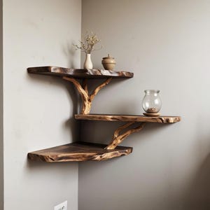 Könnte beinhalten: Ein dreistufiges Eckregal aus Naturholz mit einem lebenden Kanten-Design. Die Regalbretter werden von Ästen getragen, die an Baumäste erinnern. Das obere Regal hat eine kleine Vase mit getrockneten Blumen und einen Keramikbehälter. Das mittlere Regal hat ein Glasgefäß mit kleinen braunen Gegenständen. Das untere Regal ist leer.