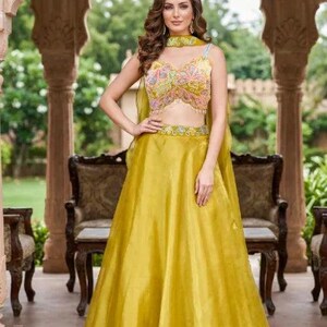 May include: A woman wearing a mustard yellow skirt and top set. The top is a cropped style with colorful floral embroidery. The skirt is a full, flowing design. The outfit is accessorized with a matching choker. The background features a classical architectural setting.