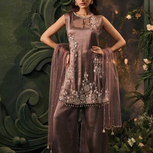May include: A mauve satin outfit, comprising a sleeveless tunic and wide-leg trousers, adorned with floral embroidery. A sheer scarf completes the look. The model is wearing the outfit in front of a green backdrop.