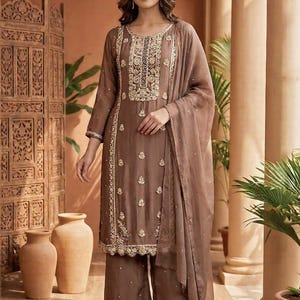 May include: A brown embroidered salwar kameez set with a matching dupatta. The outfit features gold embroidery on the neckline, sleeves, and hem. The ensemble includes wide-leg trousers and is worn with clear sandals.
