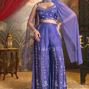 May include: A two-piece, periwinkle-colored outfit with floral embroidery. The set includes a crop top with a jeweled neckline and matching wide-leg pants. A sheer cape drapes over the shoulders. The text "Label 4U" is visible on the pants.